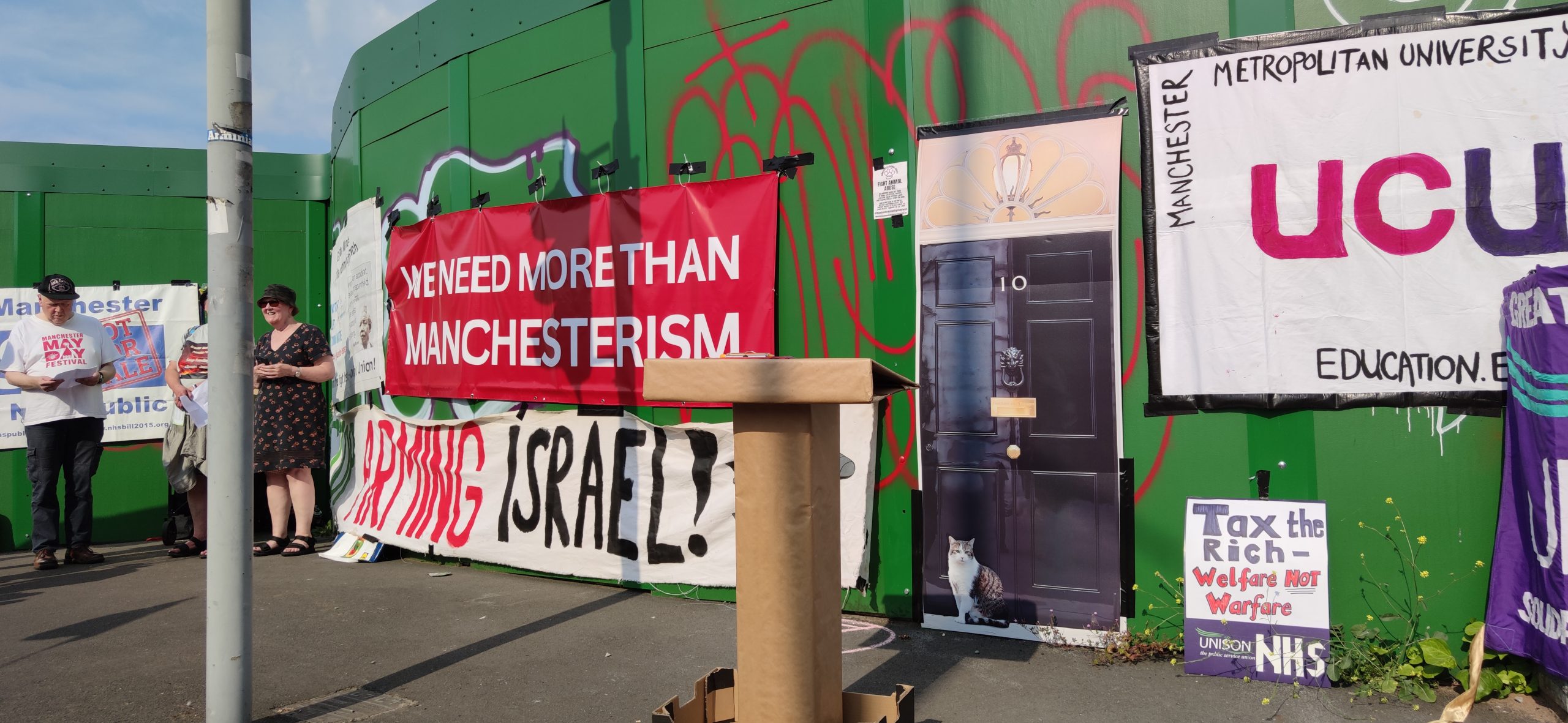 Lectern in front of replica Downing Street door, with Larry the cat, and a banner reading "We Need More Than Manchesterism"